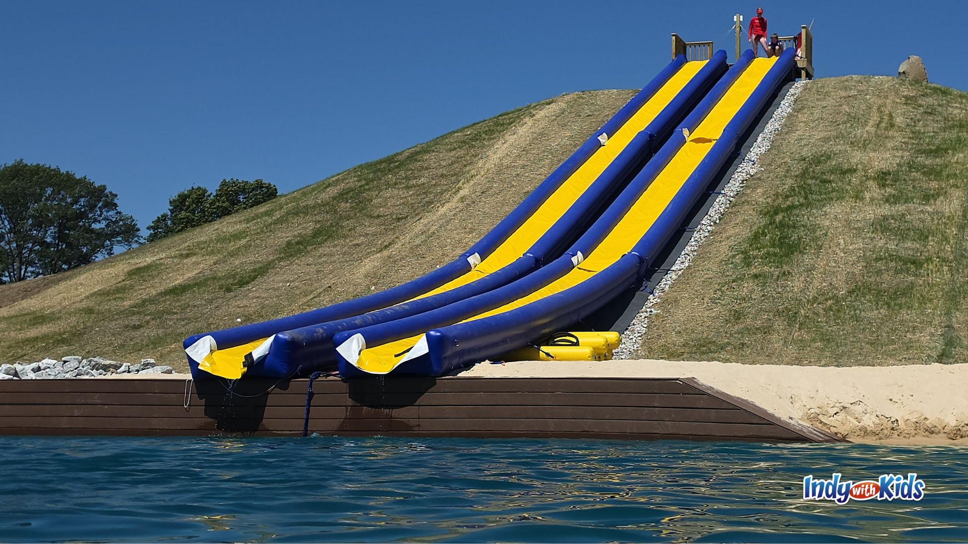A tall blue and yellow inflatable slide stretches down a grassy hill into the water at SKYLAKE Adventure Park.