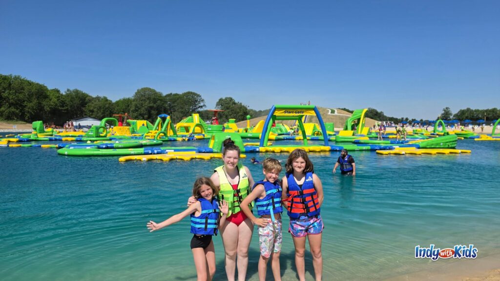 Four kids wearing life jackets smile in the shallow, clear blue water at SKYLAKE Adventure Park with inflatable obstacle courses behind them.
