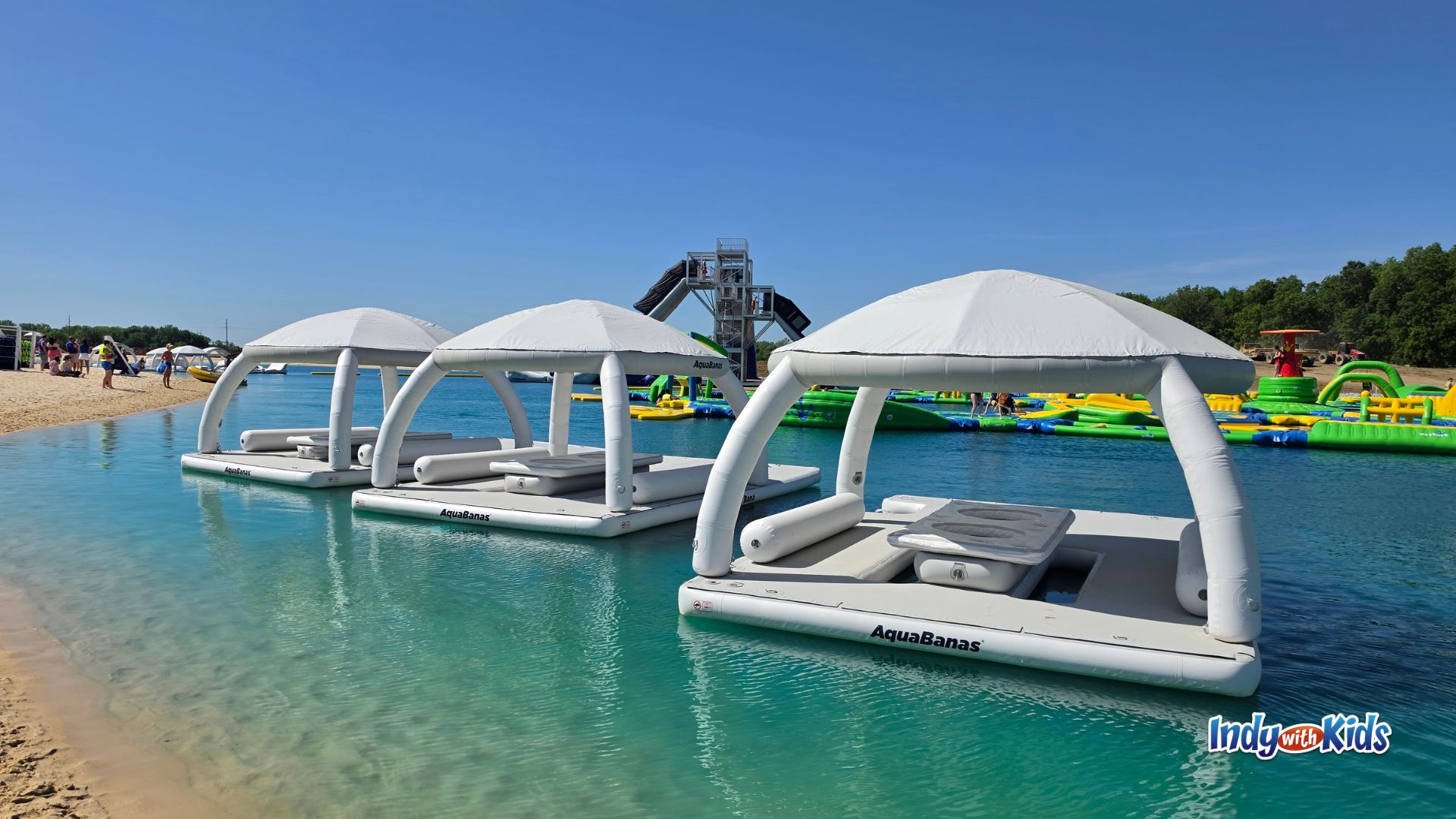 White floating Aquabana cabanas sit on the clear blue lake at SKYLAKE Adventure Park with water obstacles and the X Tower in the background.