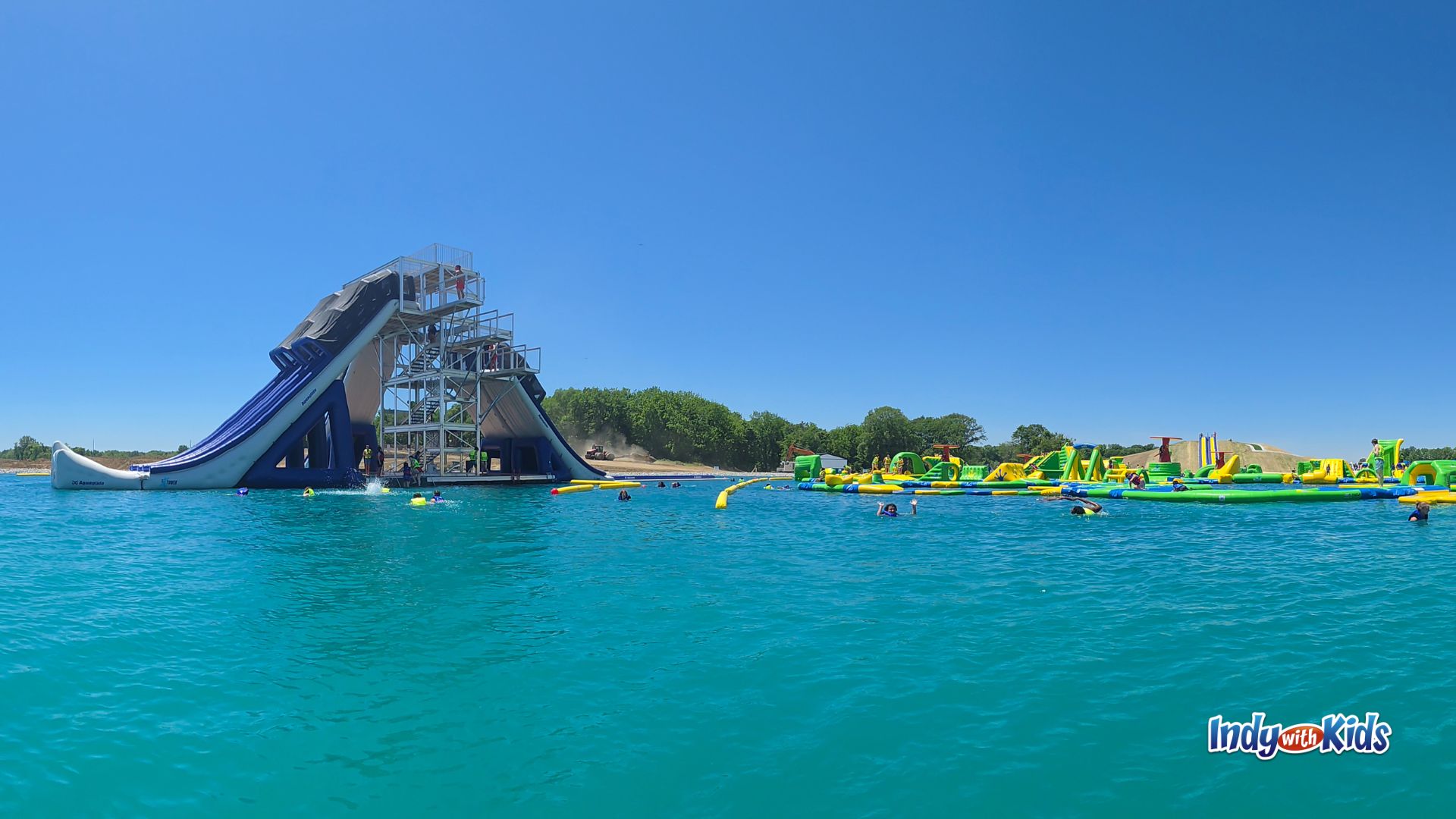 Wide view of SKYLAKE Adventure Park’s bright blue lake, with the tall X Tower slide on one side and inflatable obstacle courses across the water.