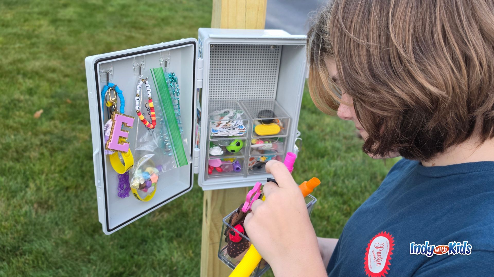 A child looks inside an open trinket trade box filled with keychains, bracelets, small toys, stickers, and other tiny treasures.