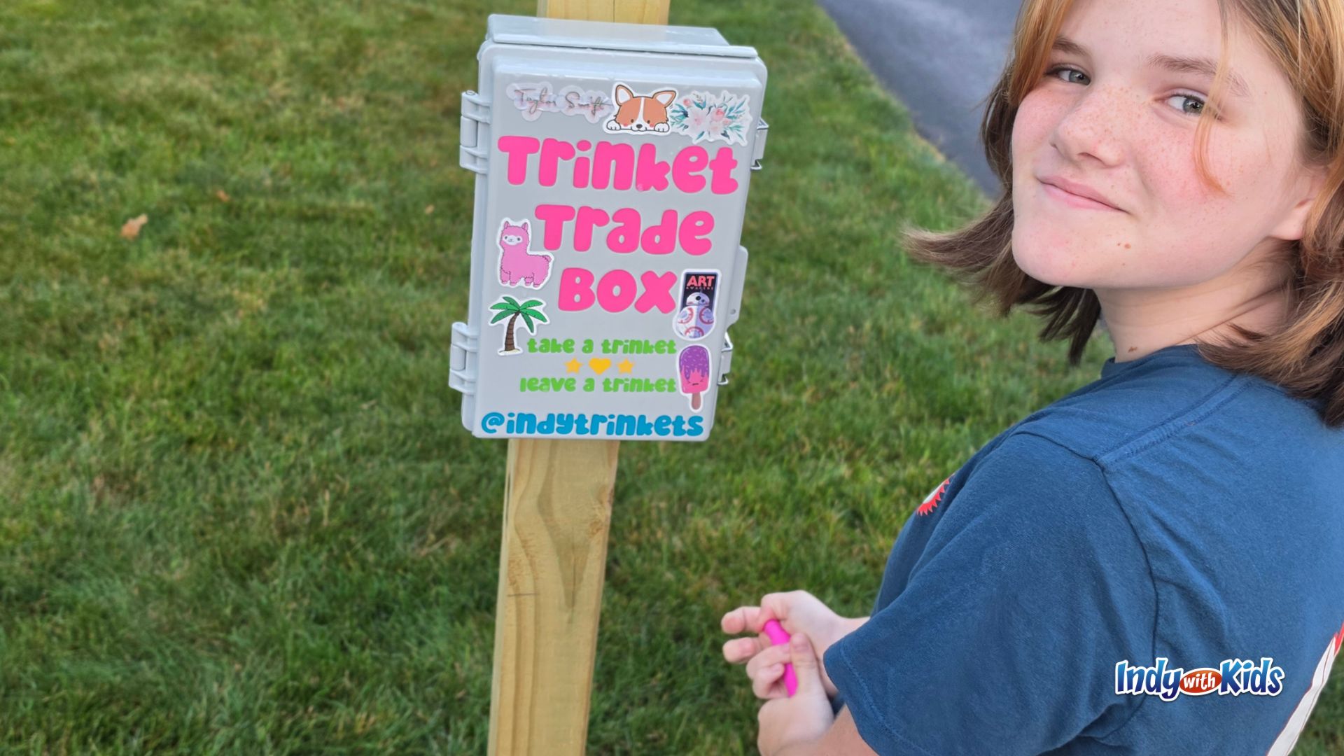 A child stands next to a small outdoor box labeled “Trinket Trade Box” with instructions to take a trinket and leave a trinket.