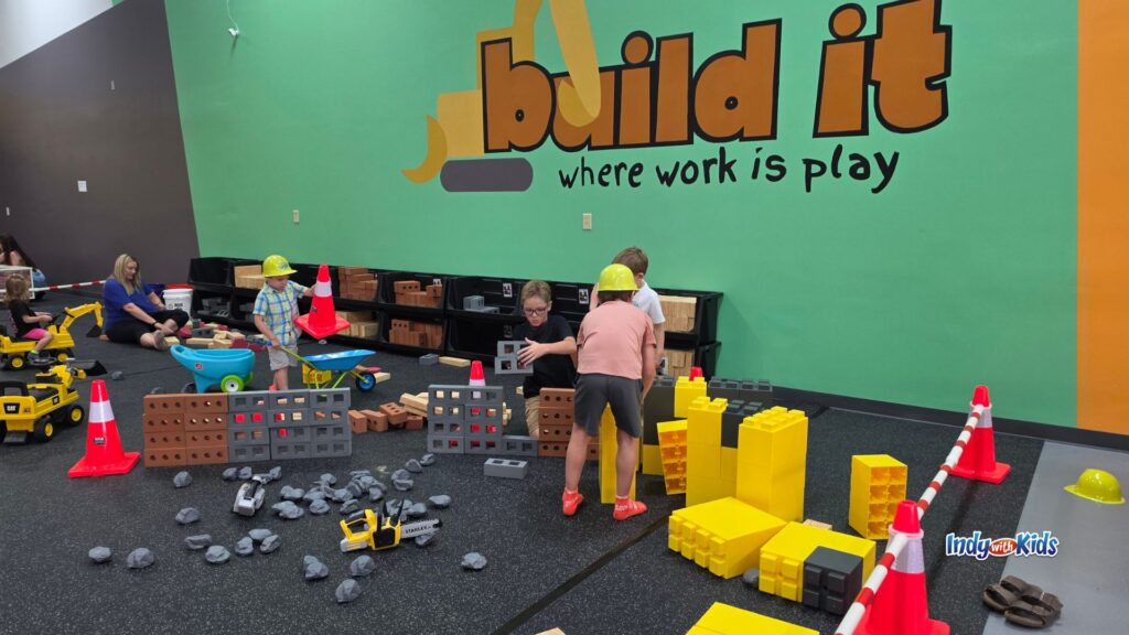 Children wearing yellow construction hats build with oversized blocks, toy bricks, cones, trucks, and pretend construction materials at Build It Playground.