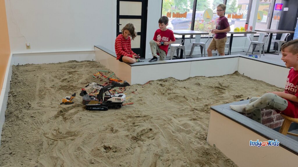 Kids play around a large indoor sand pit filled with toy excavators, trucks, and construction tools at Build It Playground.