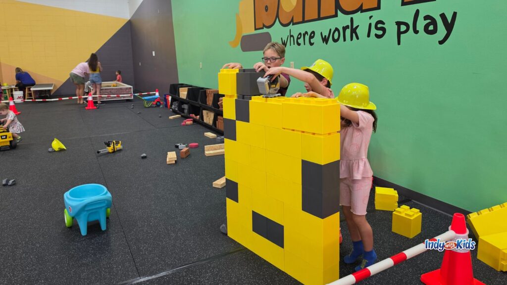 Children in yellow hard hats work together to build a tall yellow and gray structure with oversized foam-style blocks.