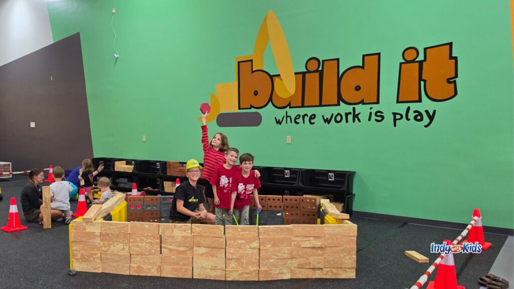 Four kids pose behind a structure they built with large wooden-style blocks under the Build It sign.