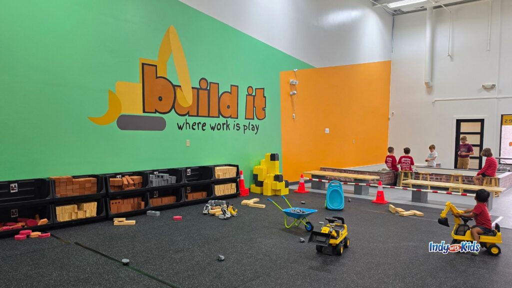 A large indoor construction-themed playground with shelves of pretend building materials, ride-on construction toys, cones, blocks, and sand play areas.