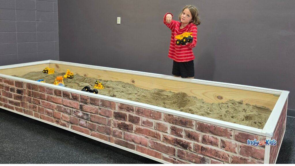 A child stands beside a large indoor sand pit, holding up a toy dump truck while construction vehicles sit in the sand.