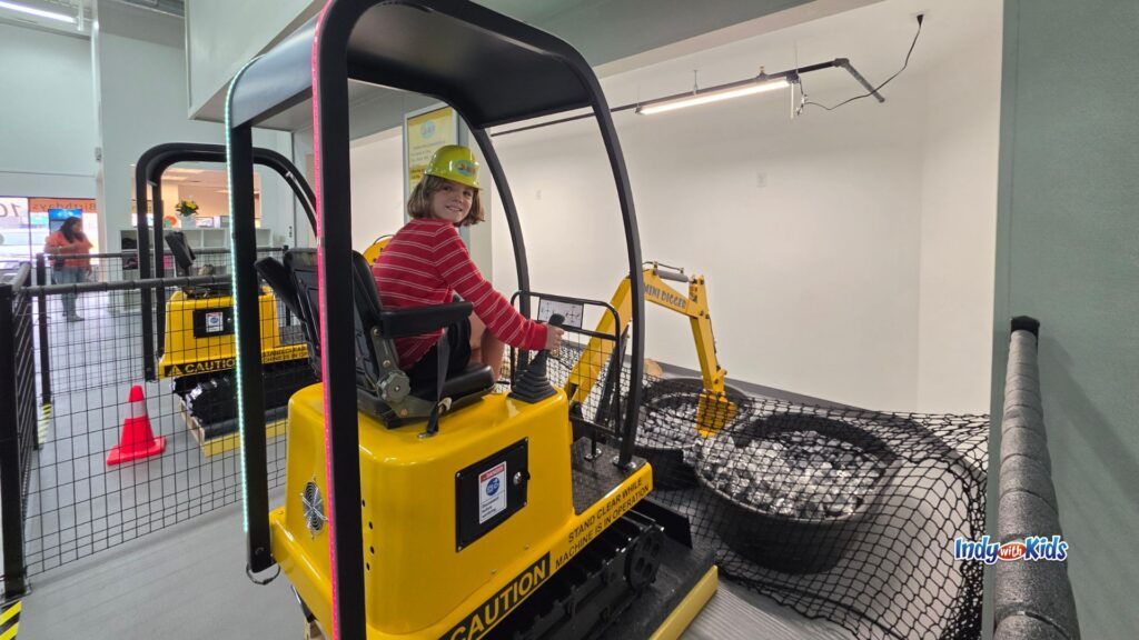 A child wearing a yellow construction helmet sits on a yellow mini excavator inside an indoor play area.