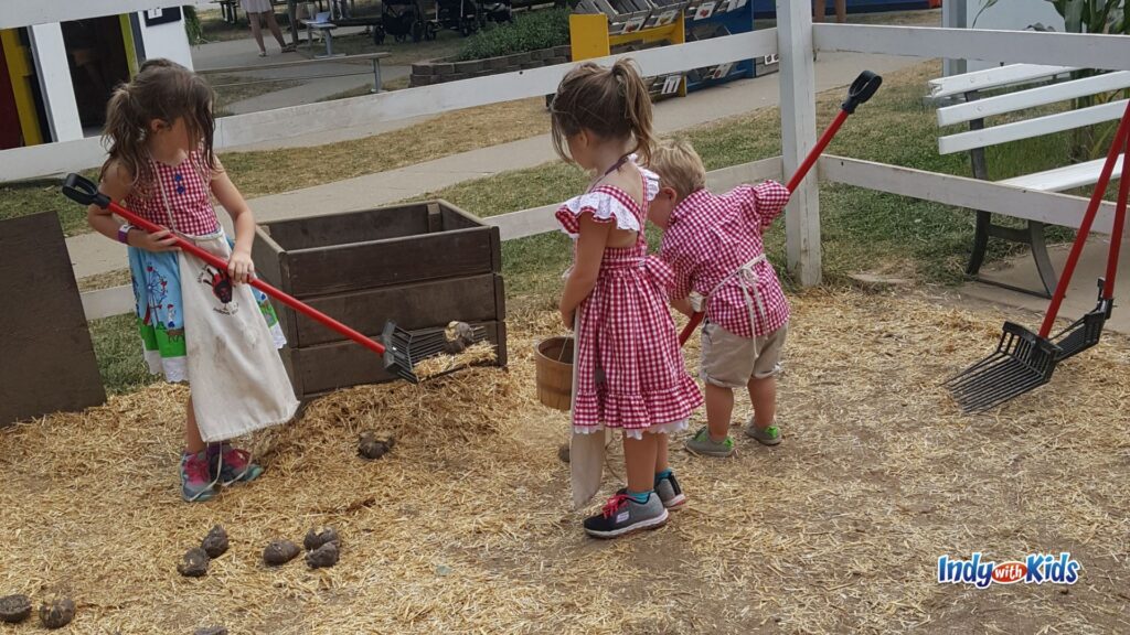 Children in red gingham outfits use small rakes and baskets in a farm pen area at Little Hands on the Farm.