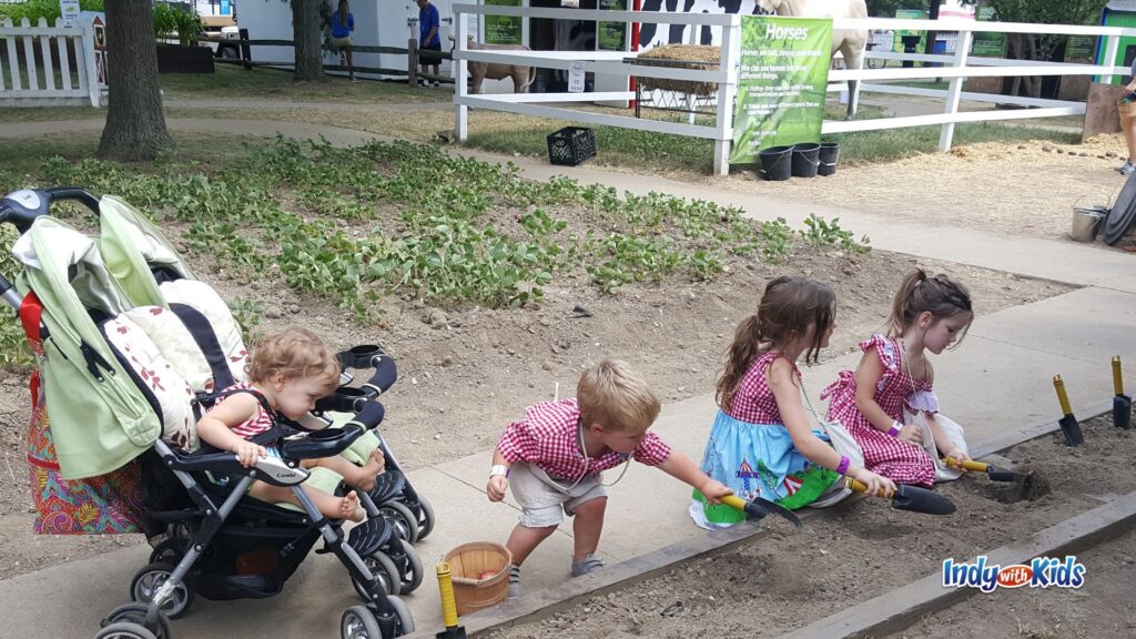 Several young children use small shovels to dig in a garden bed at Little Hands on the Farm while a baby watches from a stroller.