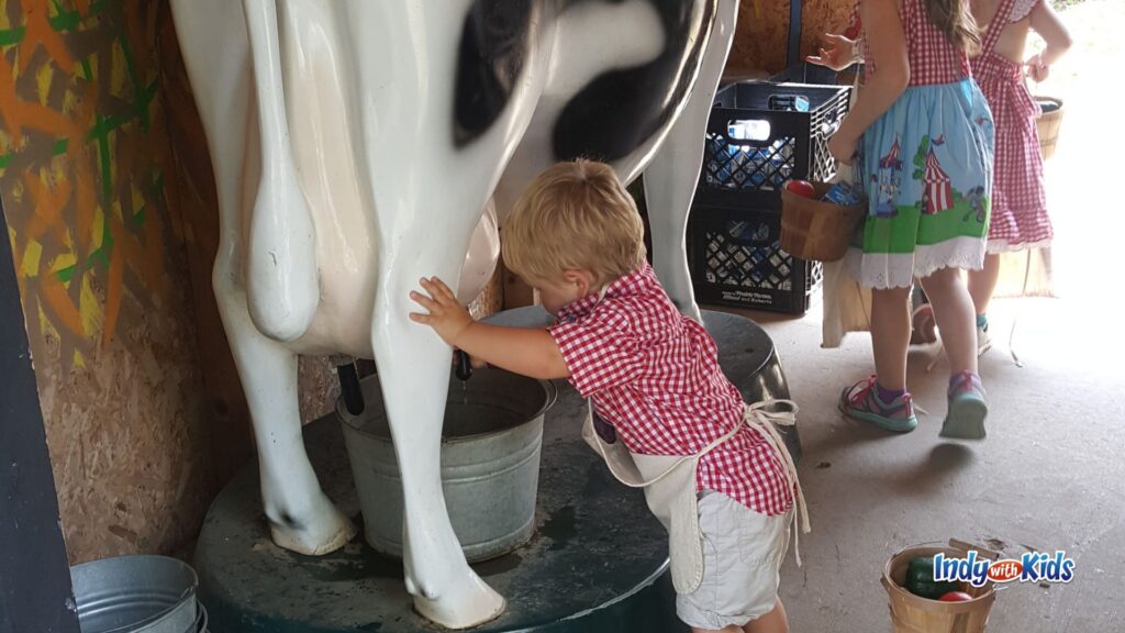 A toddler in a red gingham shirt and apron pretends to milk a cow at the Little Hands on the Farm exhibit while other children wait nearby with baskets.