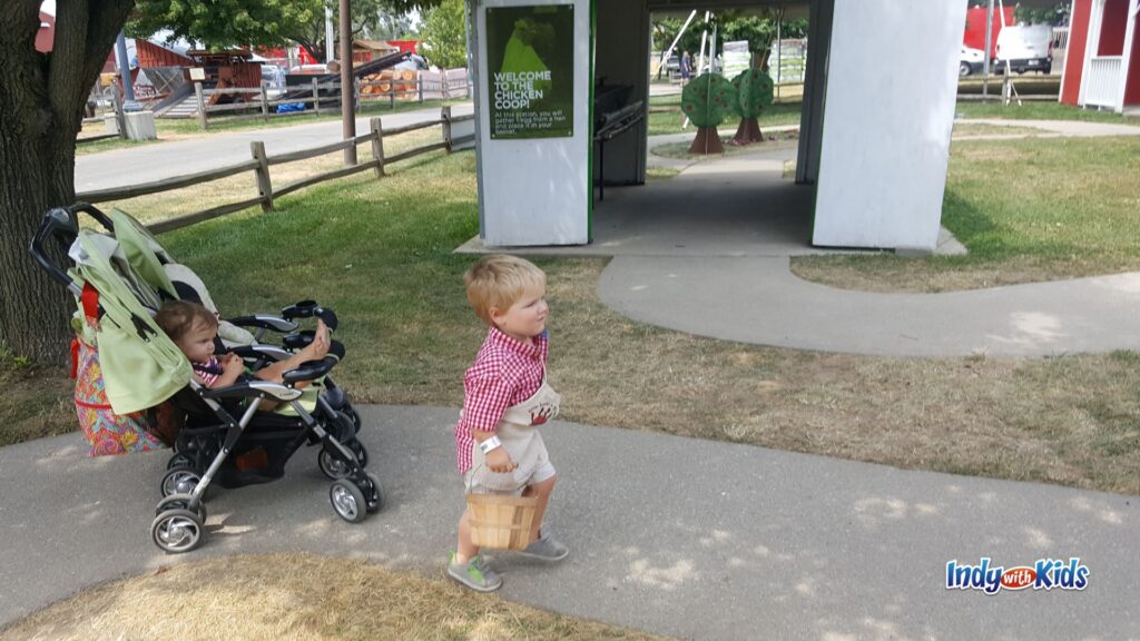 A young child carries a small wooden basket past the chicken coop station at Little Hands on the Farm while another child rides in a stroller.
